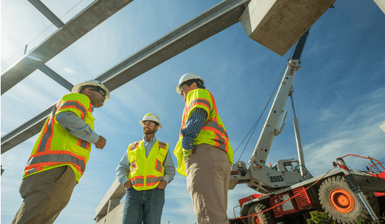 Three workers in white hard hats and safety vests on project site, seen from below